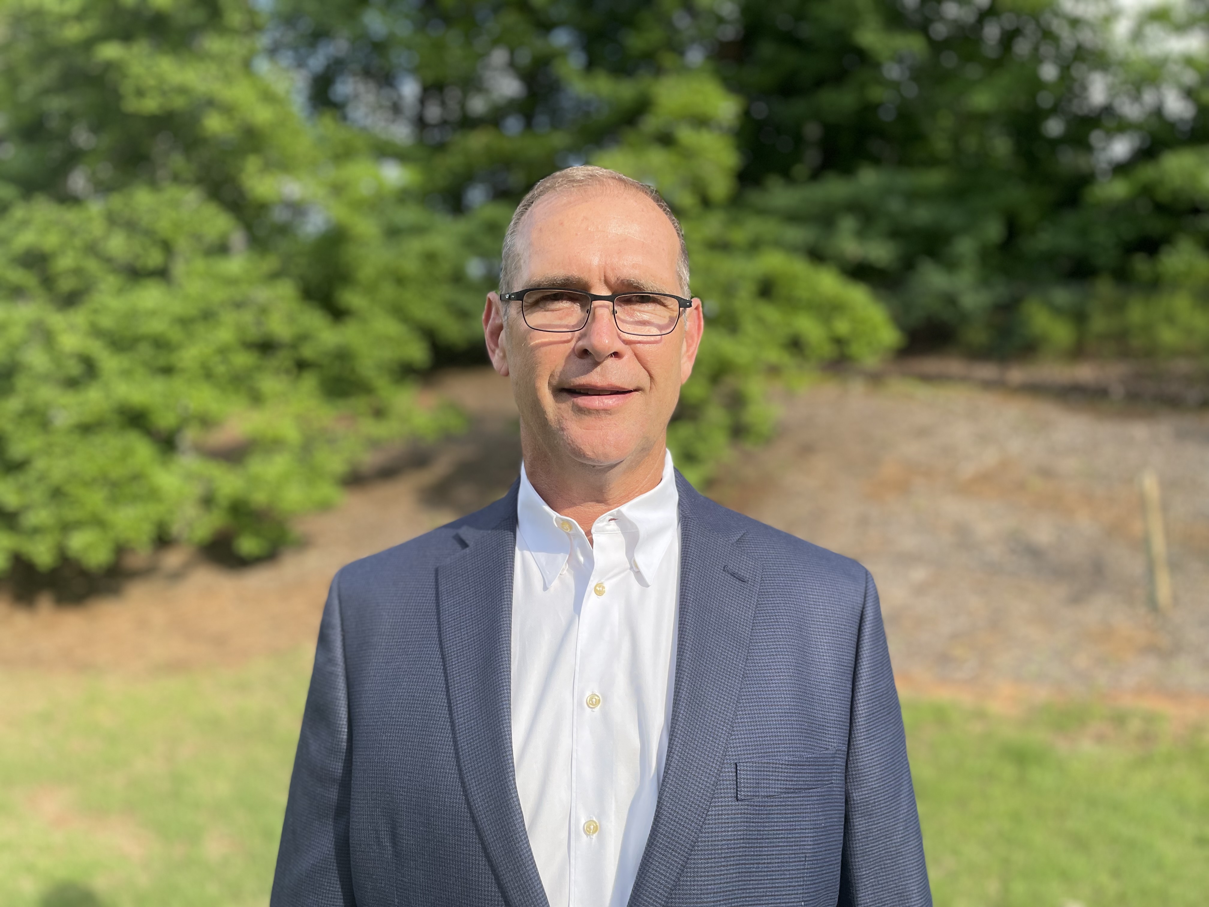 Professional in gray suit standing in green outdoor setting with trees Headshot