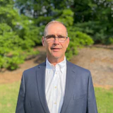 Professional in gray suit standing in green outdoor setting with trees