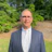 Professional in gray suit standing in green outdoor setting with trees Headshot