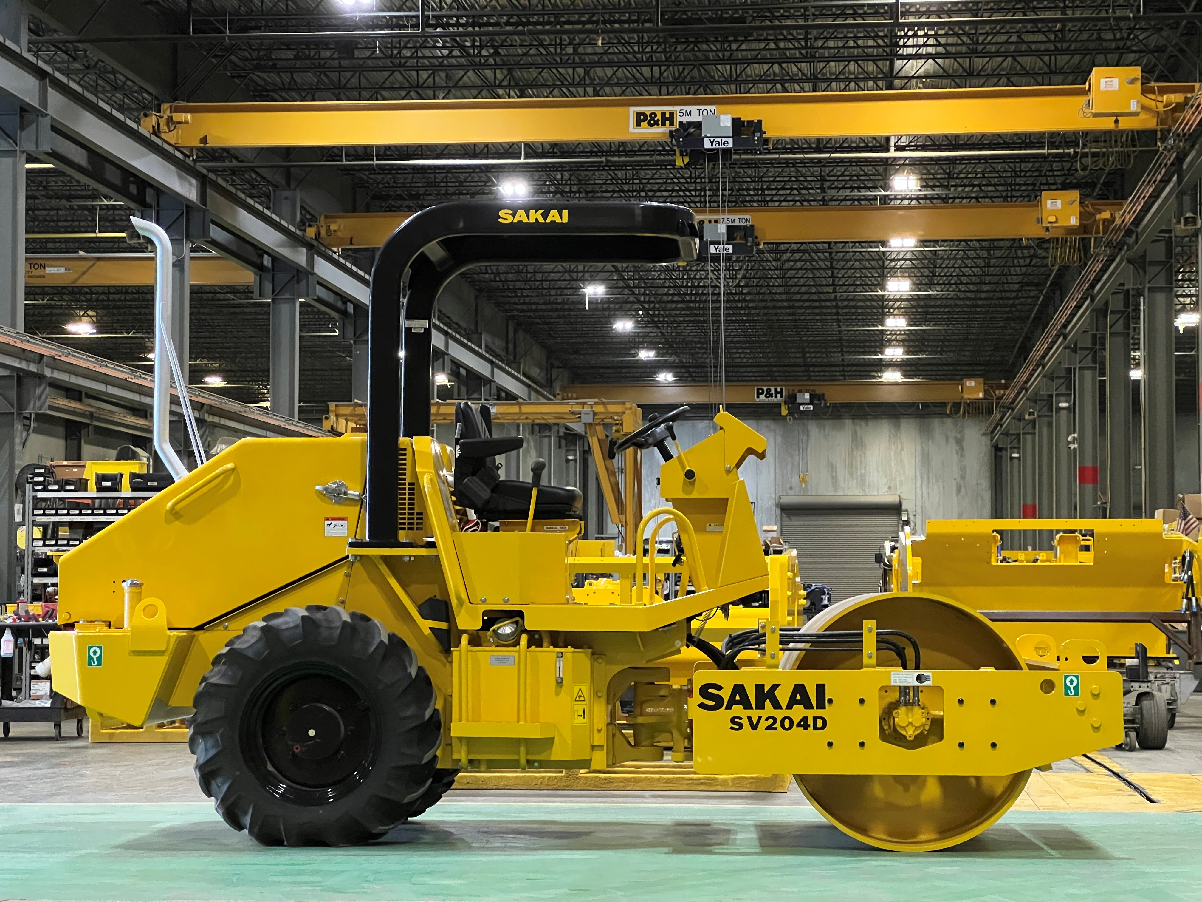 Yellow Sakai industrial road roller in manufacturing facility with overhead crane