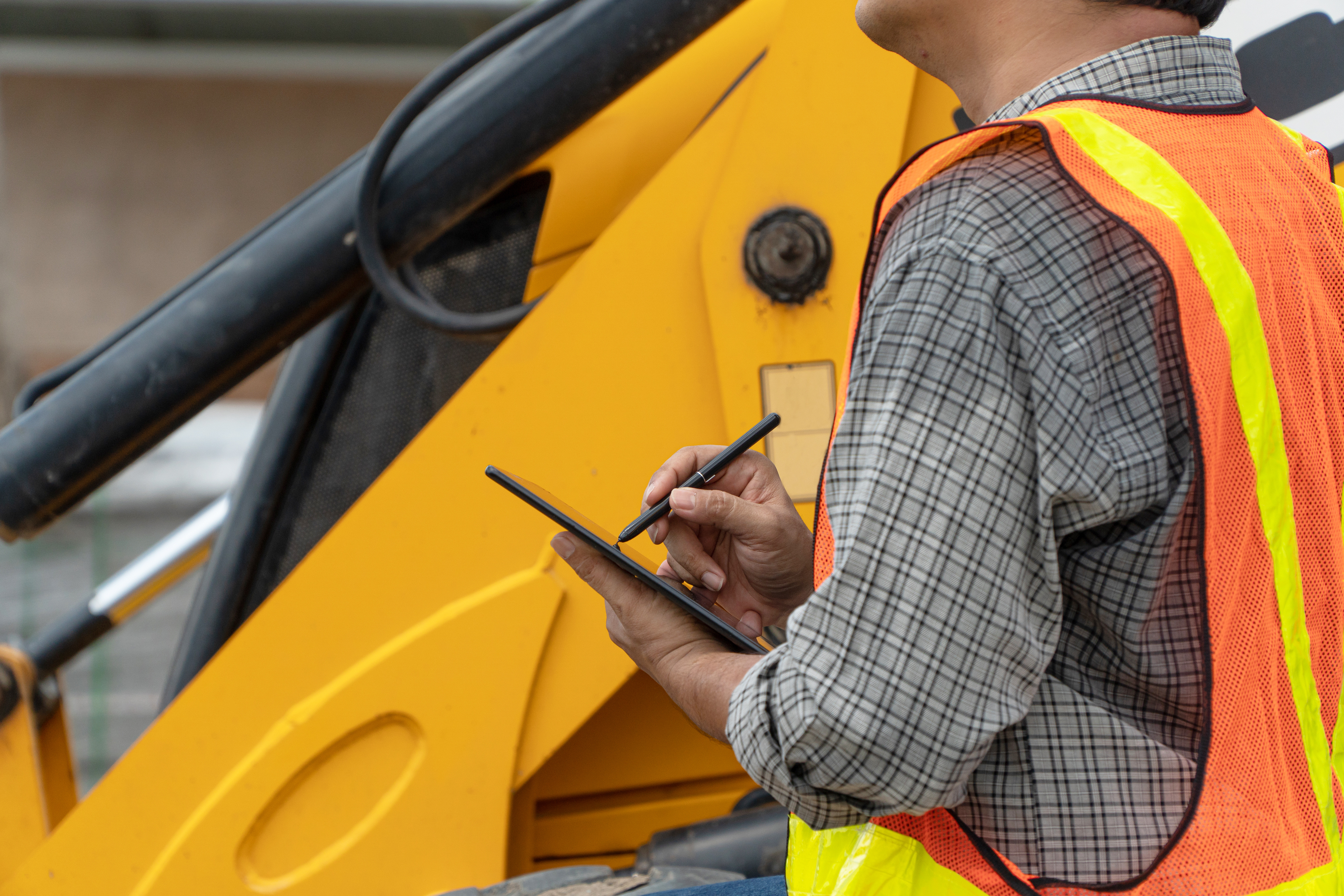 Worker in safety vest using tablet near yellow construction equipment