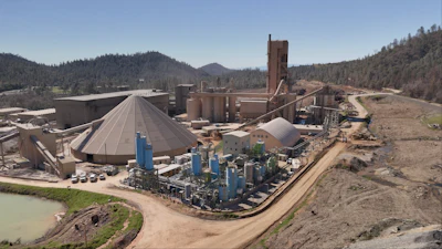 Industrial processing facility with blue tanks and machinery in mountainous forest landscape