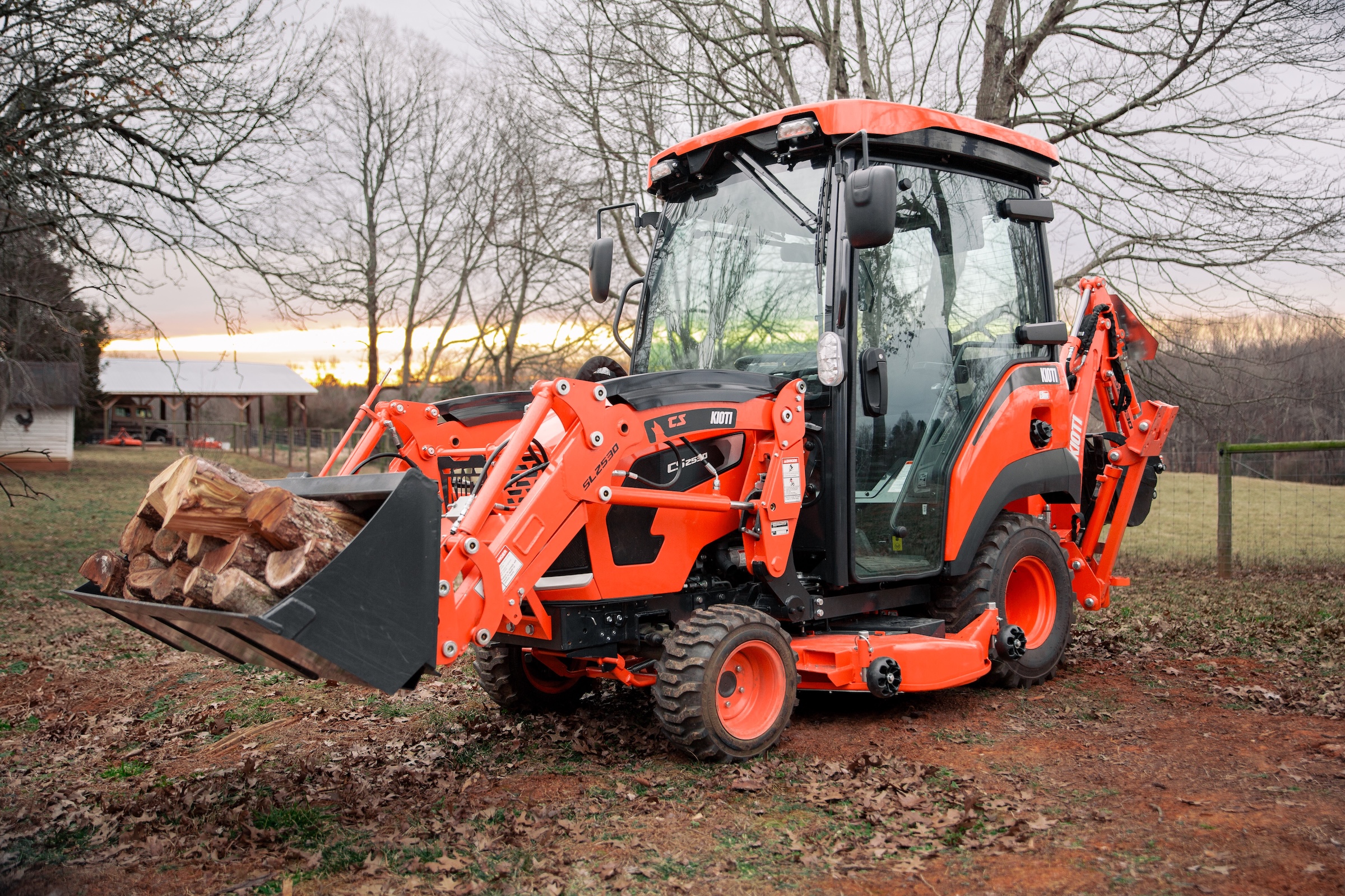KIOTI CS30 sub-compact tractor with front loader carrying firewood logs in rural setting