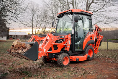 KIOTI CS30 sub-compact tractor with front loader carrying firewood logs in rural setting