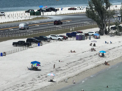 Crowded beach with parked cars, colorful umbrellas, and sandy shoreline beside highway