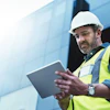 Construction worker in hard hat and safety vest using tablet near modern building