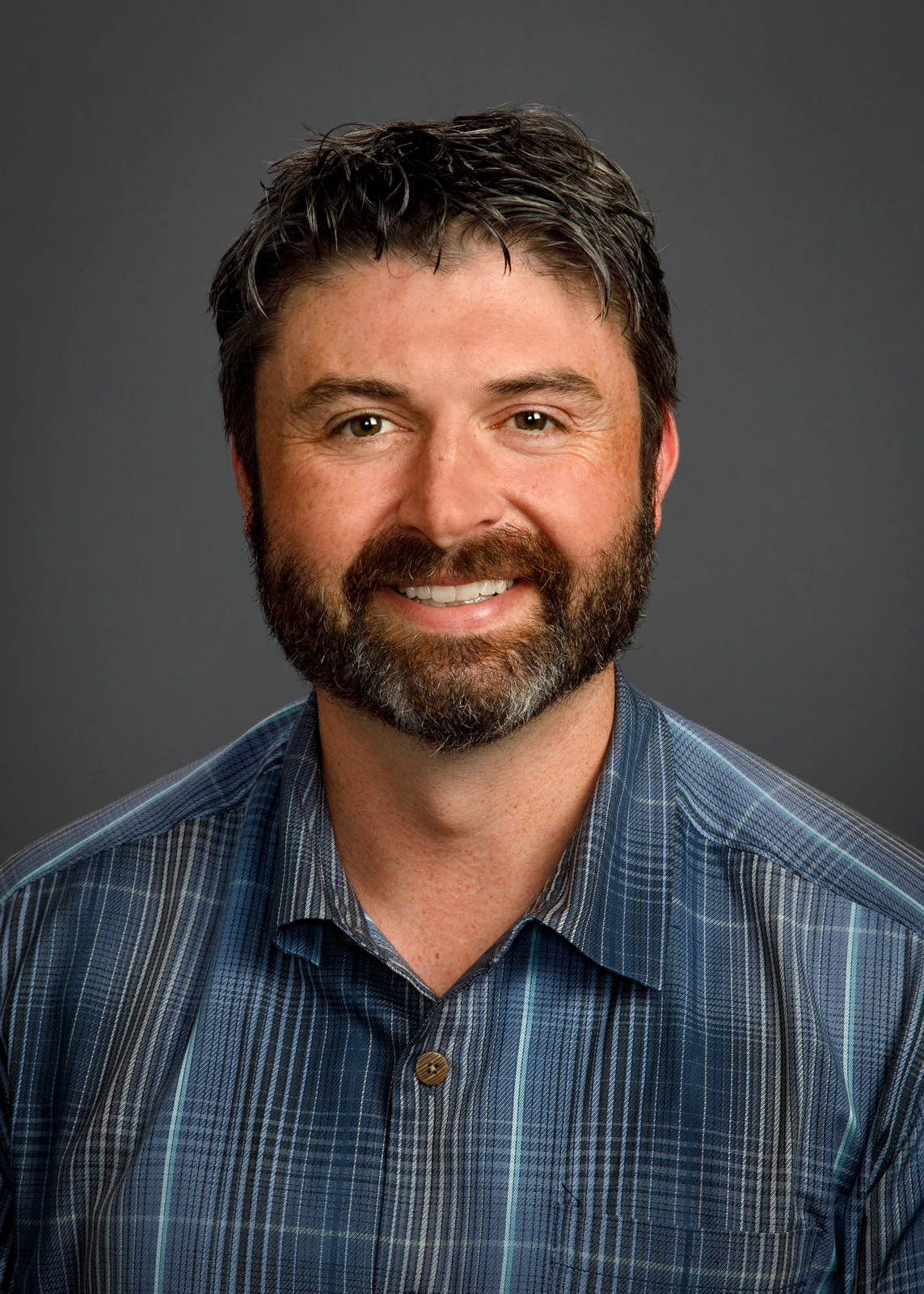 Professional headshot of smiling man with dark beard wearing blue plaid shirt against gray background Headshot