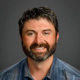 Professional headshot of smiling man with dark beard wearing blue plaid shirt against gray background
