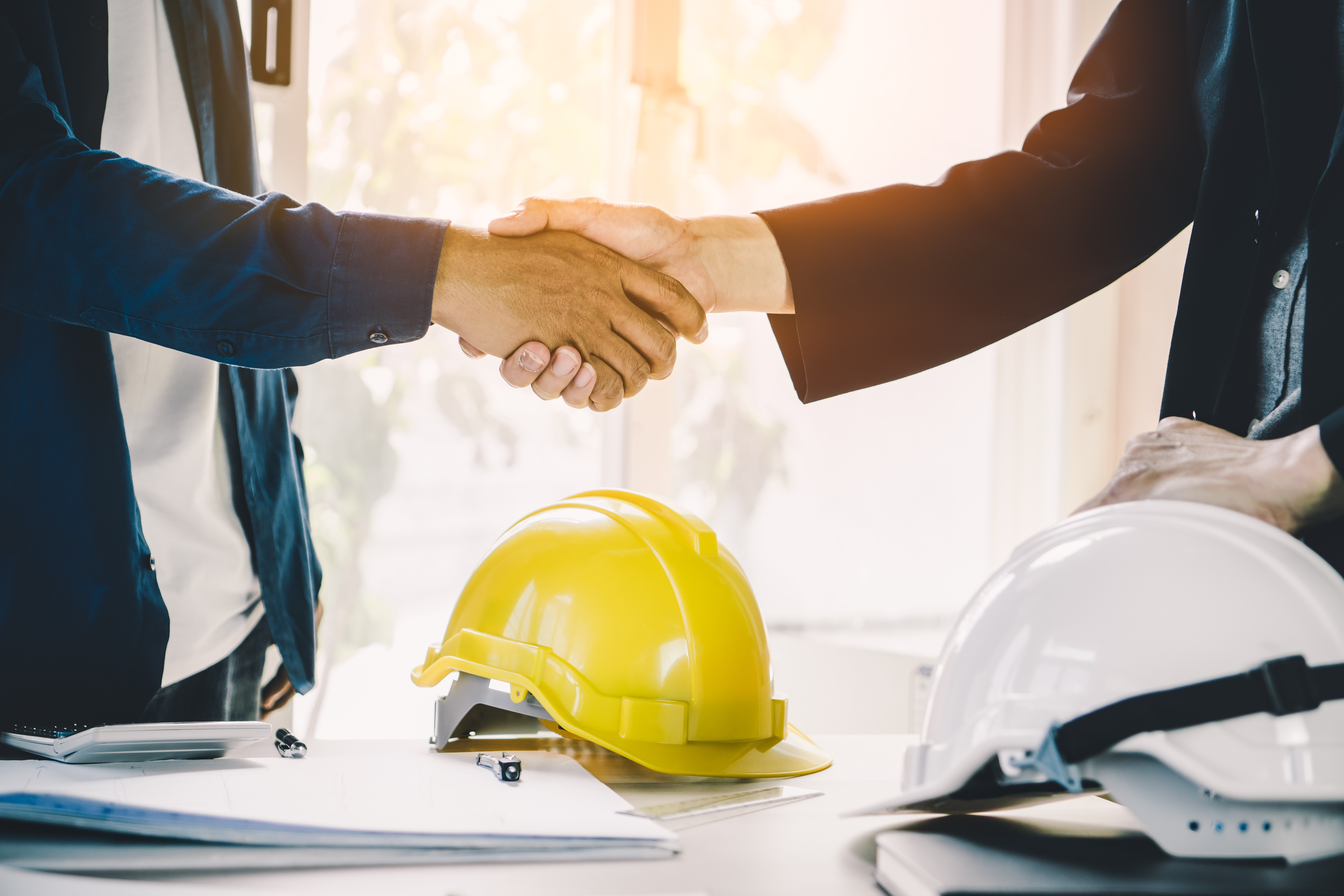 Business professionals shaking hands near yellow and white construction helmets
