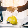 Business professionals shaking hands near yellow and white construction helmets