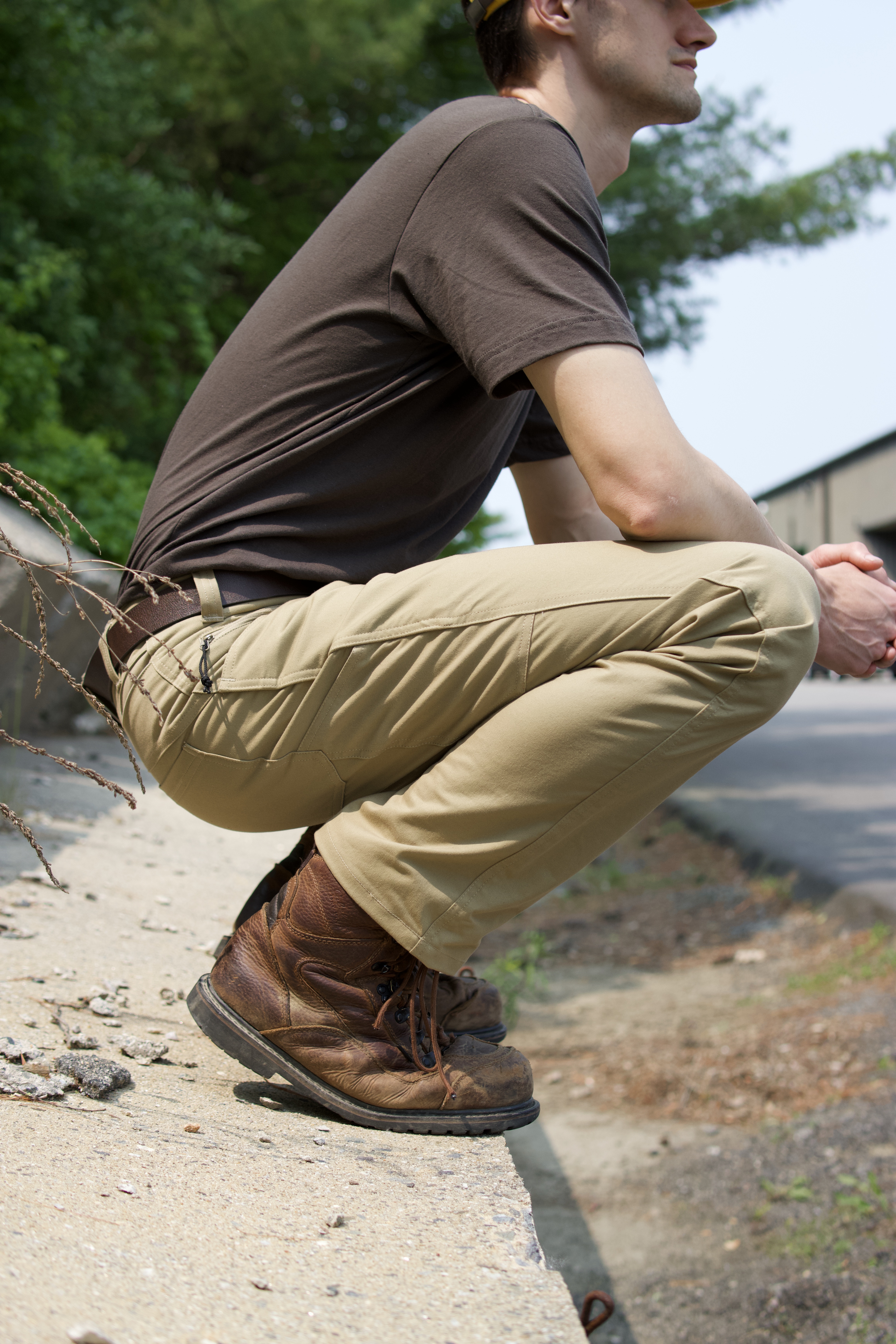 Person in brown shirt and khaki pants crouching on concrete edge near greenery