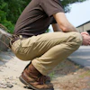 Person in brown shirt and khaki pants crouching on concrete edge near greenery