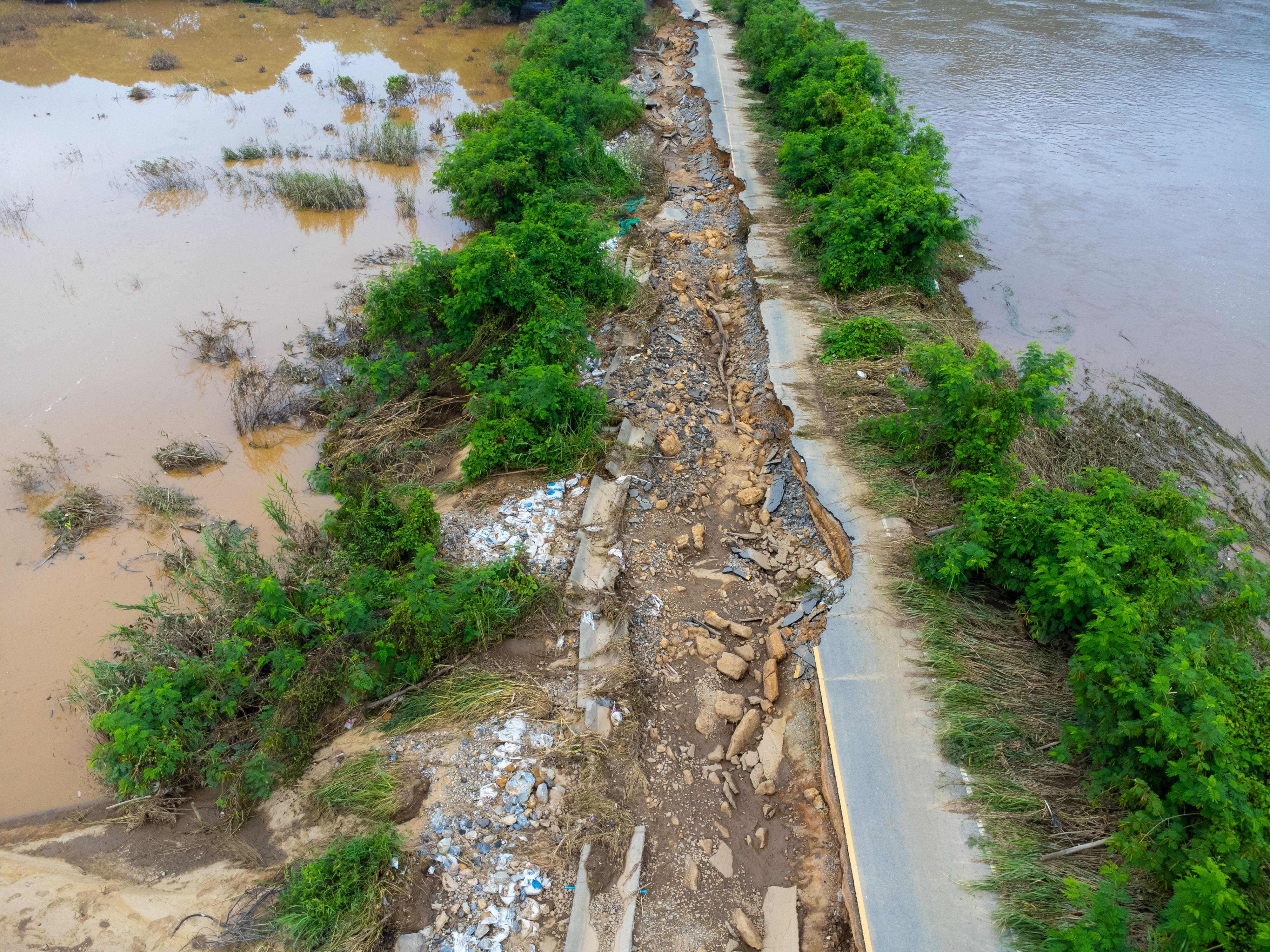 Flood-damaged road with rocky debris, surrounded by muddy water and green trees