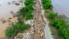 Flood-damaged road with rocky debris, surrounded by muddy water and green trees