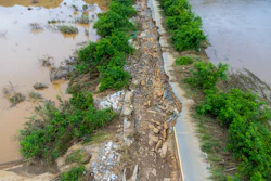 Flood-damaged road with rocky debris, surrounded by muddy water and green trees