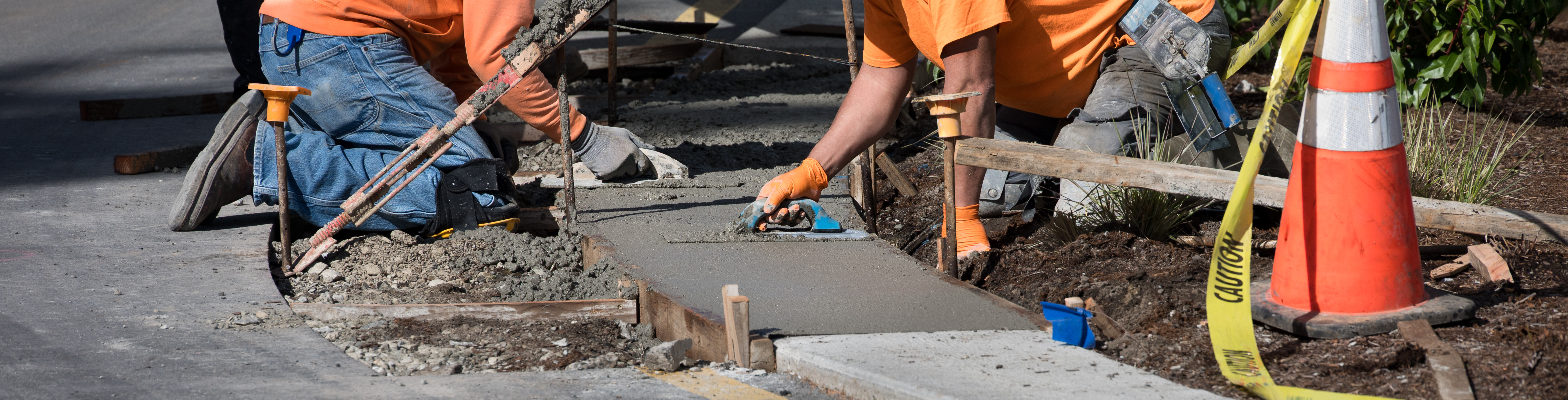 Construction workers pouring and smoothing concrete sidewalk with safety cone