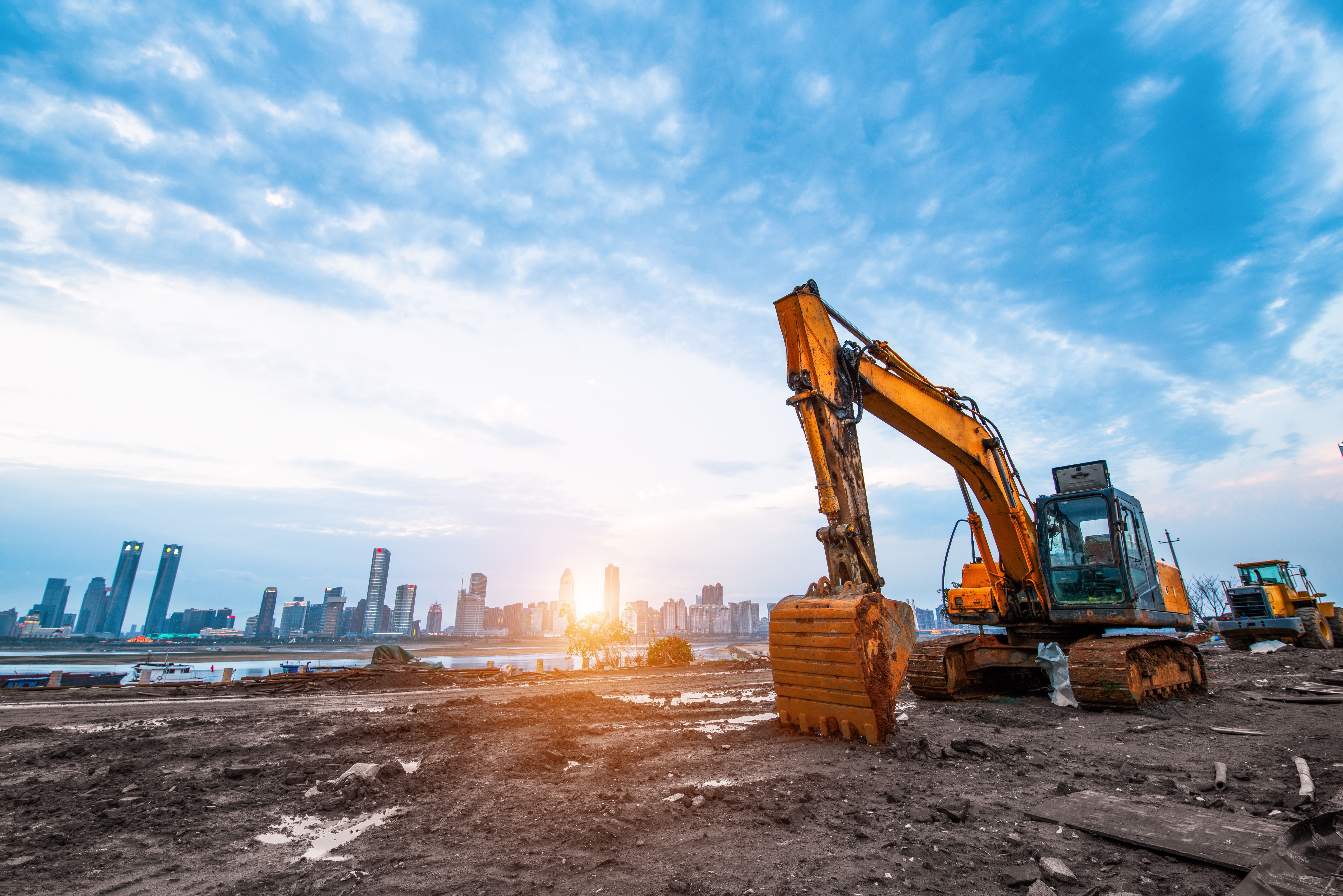 Yellow excavator on muddy construction site with urban skyline at sunrise