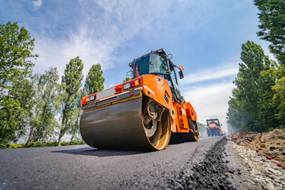 Orange road roller compacting fresh asphalt on highway with trees in background