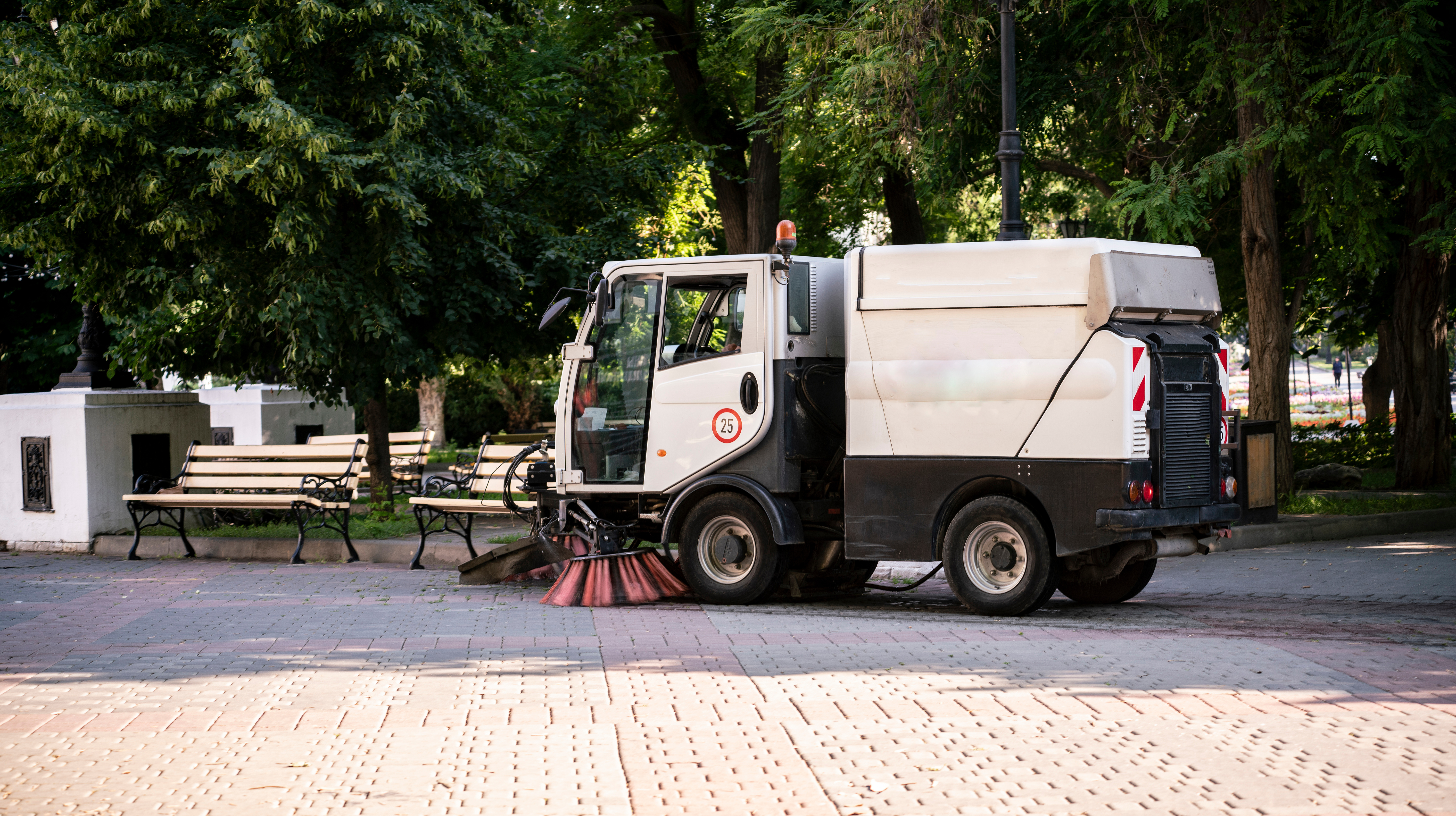 Street sweeping truck cleaning urban park pathway with trees and benches
