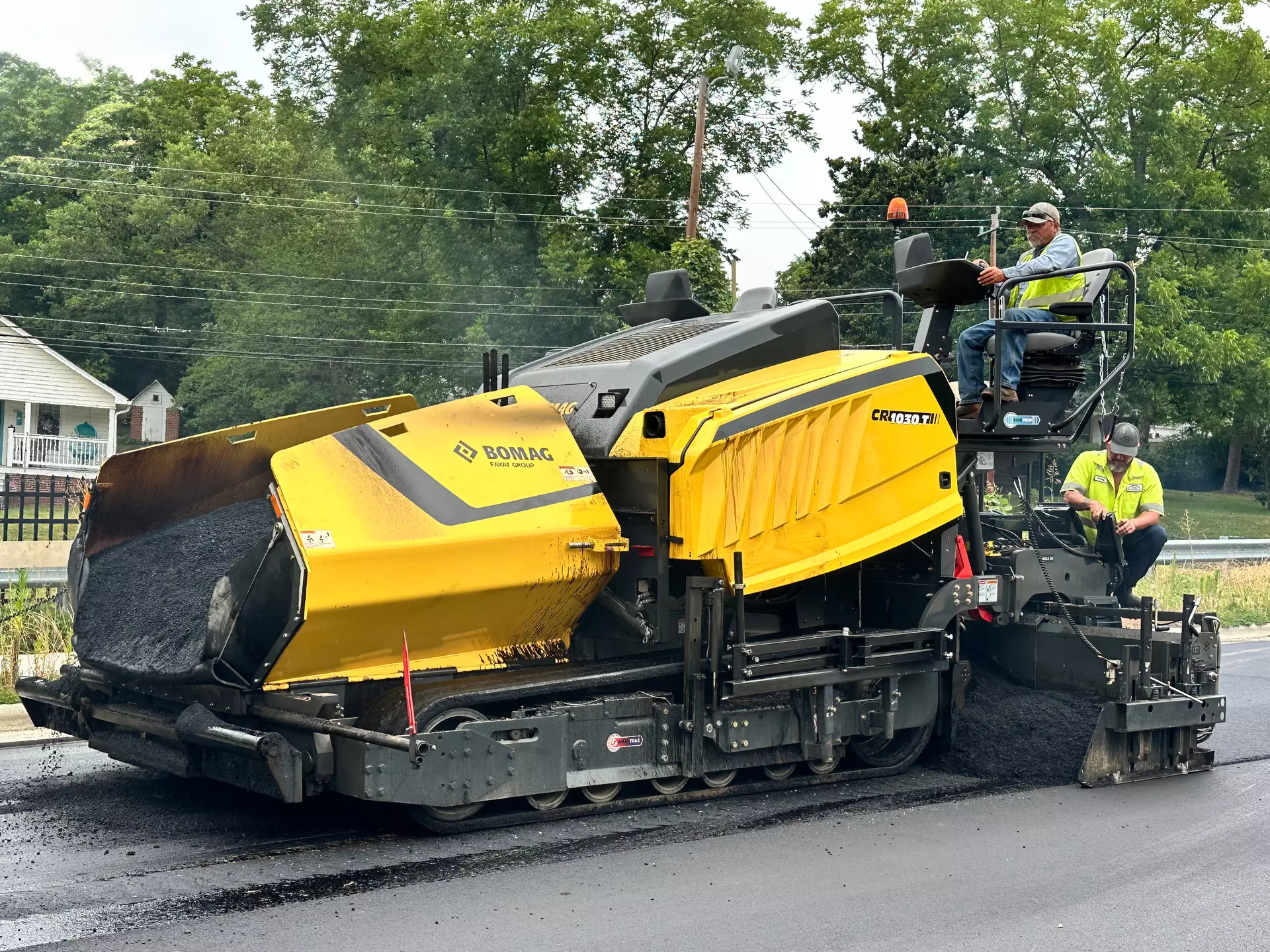 Yellow Bomag road paving machine spreading fresh asphalt on street with workers
