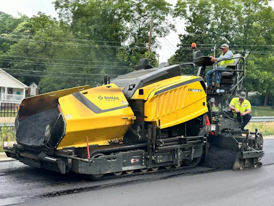 Yellow Bomag road paving machine spreading fresh asphalt on street with workers