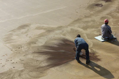 Two workers on sandy ground, one crouching and one sitting, creating sweeping patterns