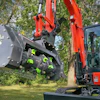 Red compact excavator with forestry mulching attachment clearing vegetation near trees