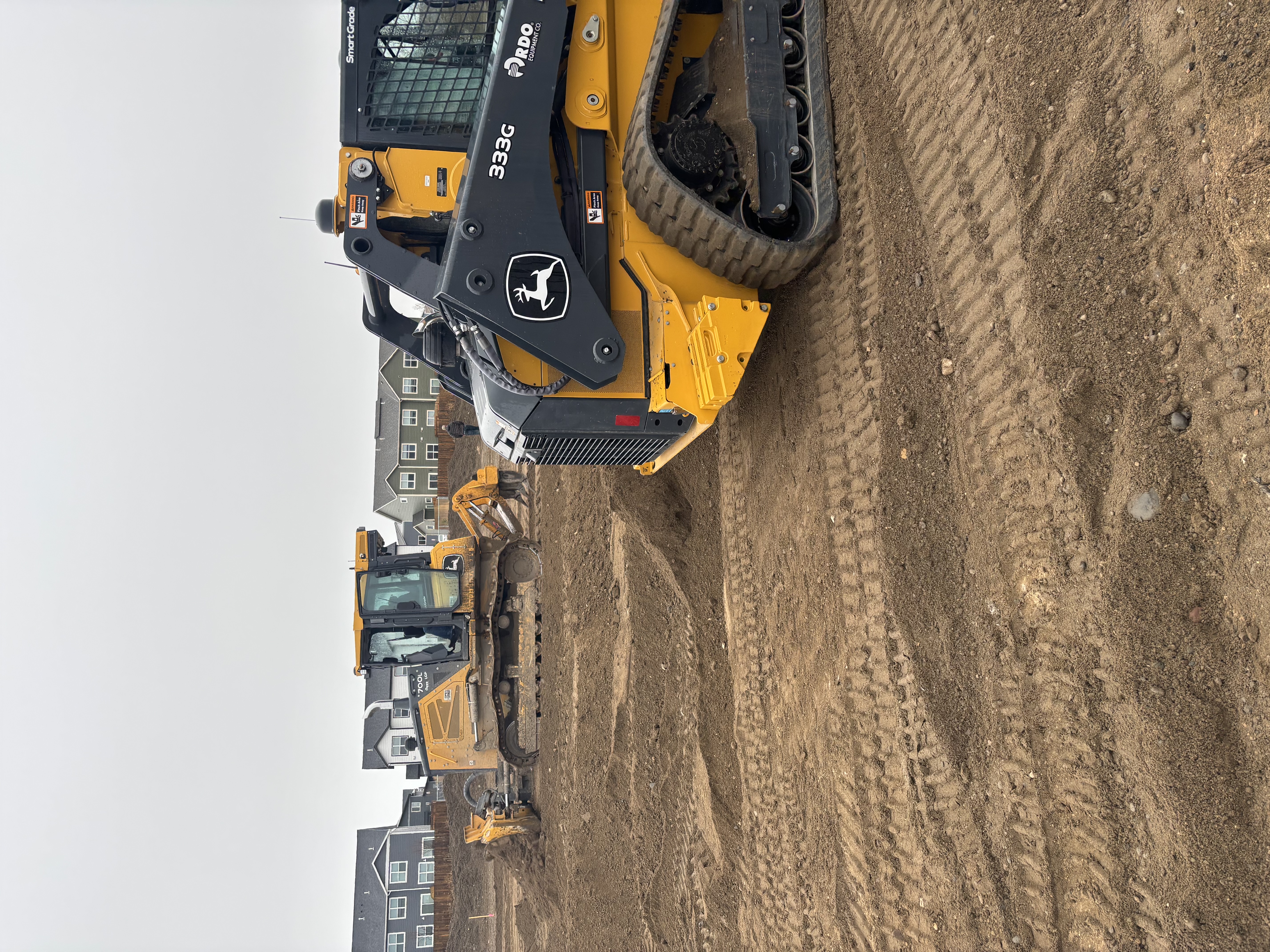Yellow John Deere construction vehicles working on dirt lot near residential development