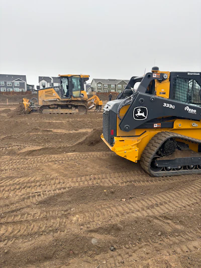 Yellow John Deere construction vehicles working on dirt lot near residential development