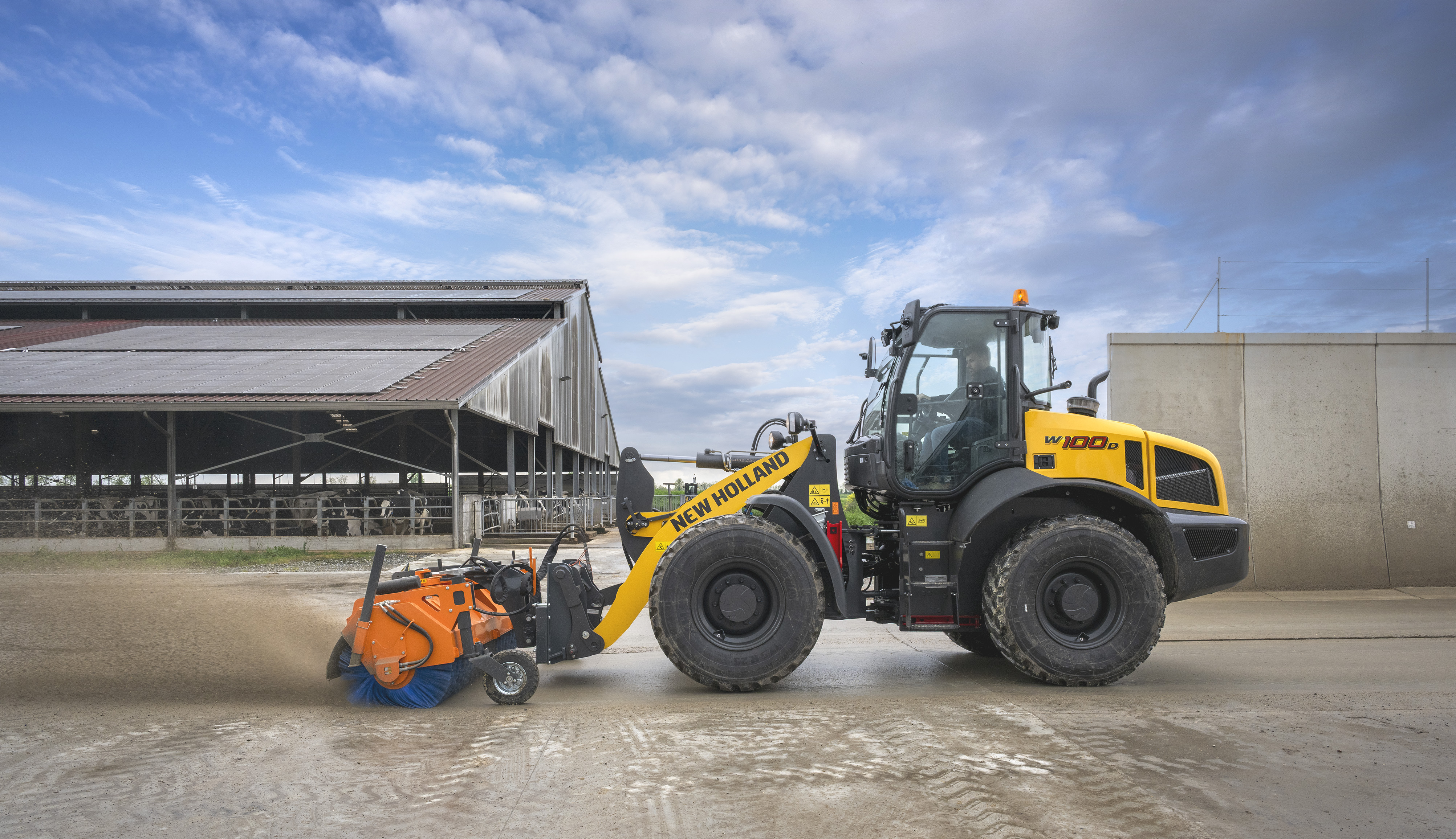 Yellow New Holland wheel loader with orange street sweeper attachment near agricultural building