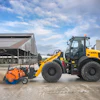 Yellow New Holland wheel loader with orange street sweeper attachment near agricultural building