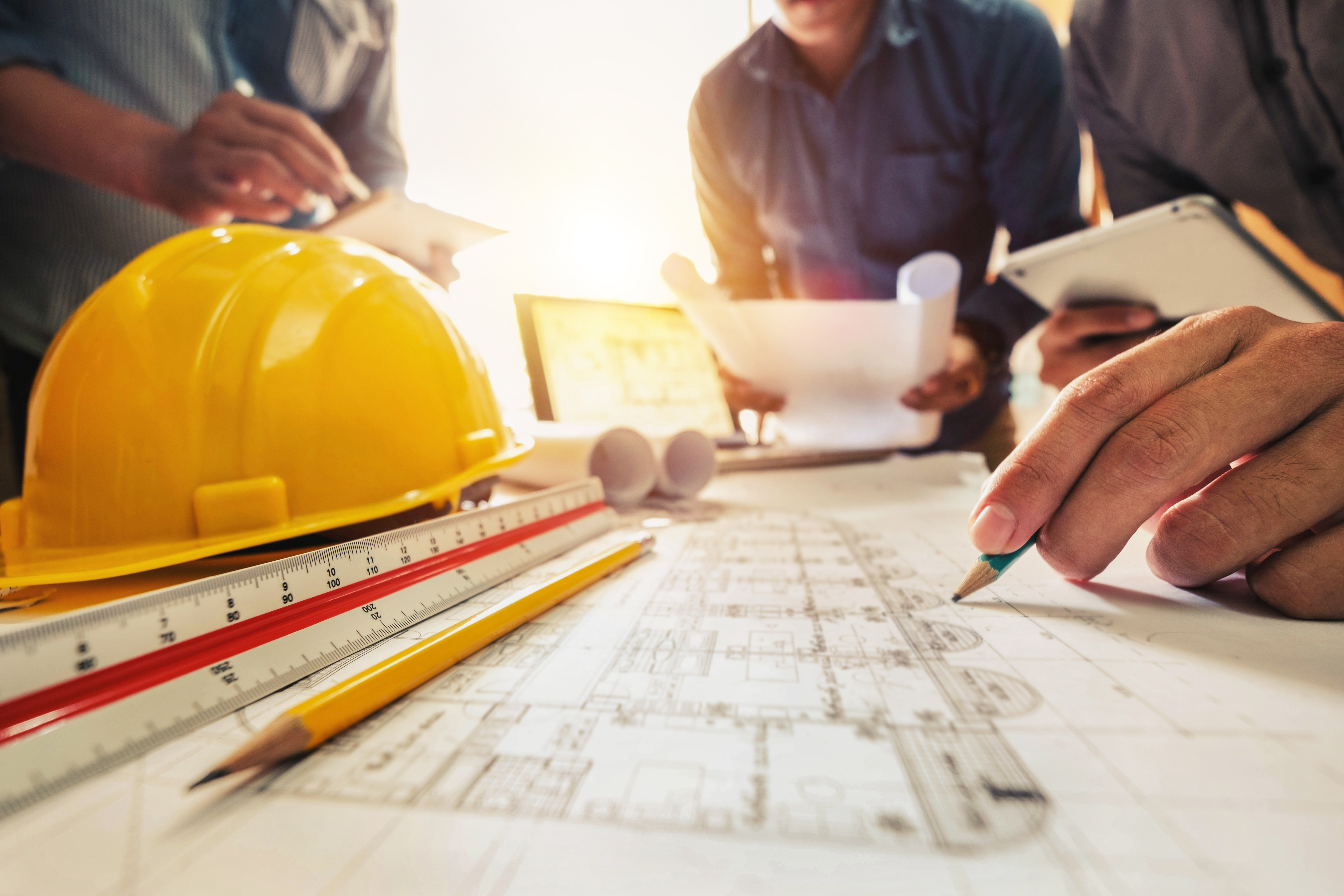 Construction blueprint with yellow hard hat, drafting tools, and hands working on architectural plans