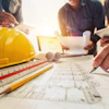 Construction blueprint with yellow hard hat, drafting tools, and hands working on architectural plans