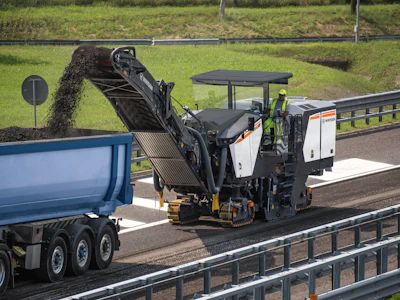 Road construction machine milling asphalt on highway with blue dump truck