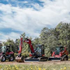 Red and gray construction vehicles lined up in grassy field with trees