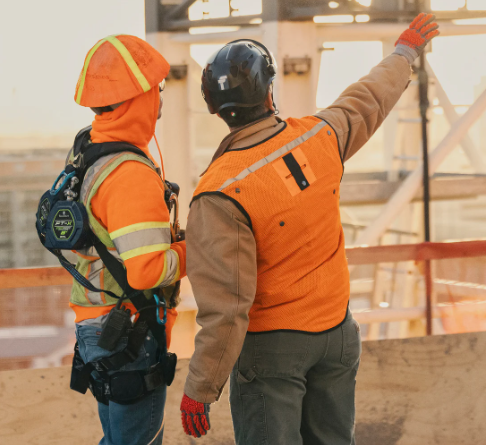 Two construction workers in orange safety vests surveying a construction site