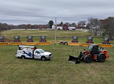 Snow plow trucks and service vehicles lined up on grassy field with flag and barns