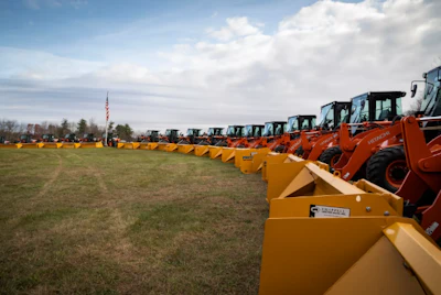 Row of orange construction loaders and excavators lined up on grassy field with flag