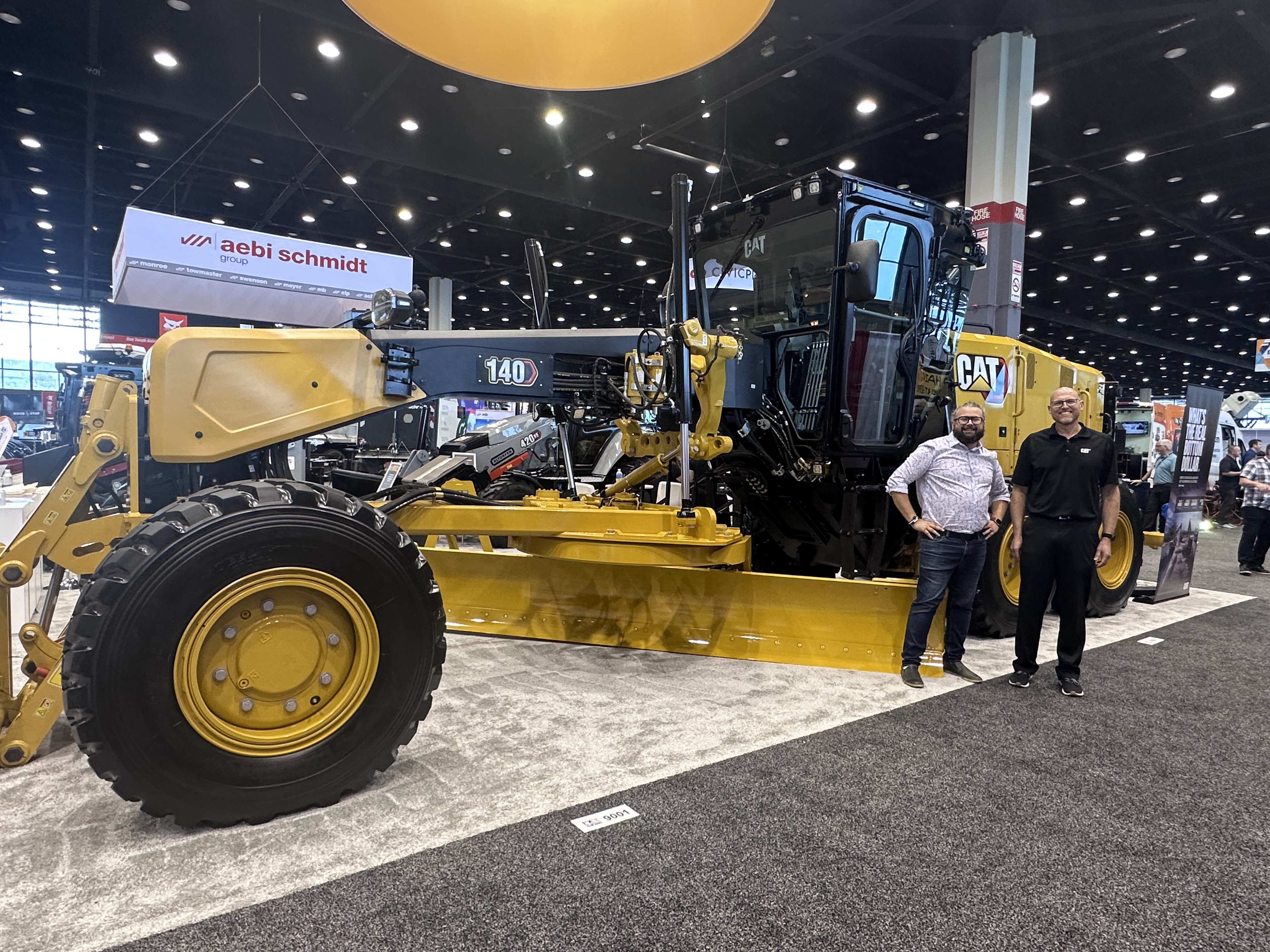 Yellow Caterpillar motor grader on display at industrial trade show with two people standing nearby