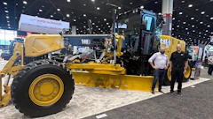Yellow Caterpillar motor grader on display at industrial trade show with two people standing nearby