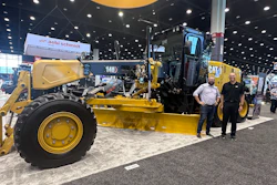 Yellow Caterpillar motor grader on display at industrial trade show with two people standing nearby
