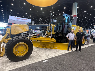 Yellow Caterpillar motor grader on display at industrial trade show with two people standing nearby