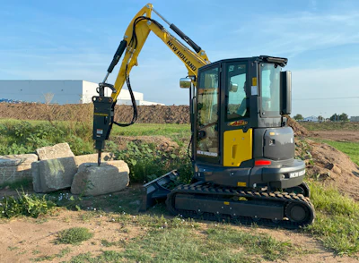 Yellow New Holland mini excavator working with large stones on grassy construction site