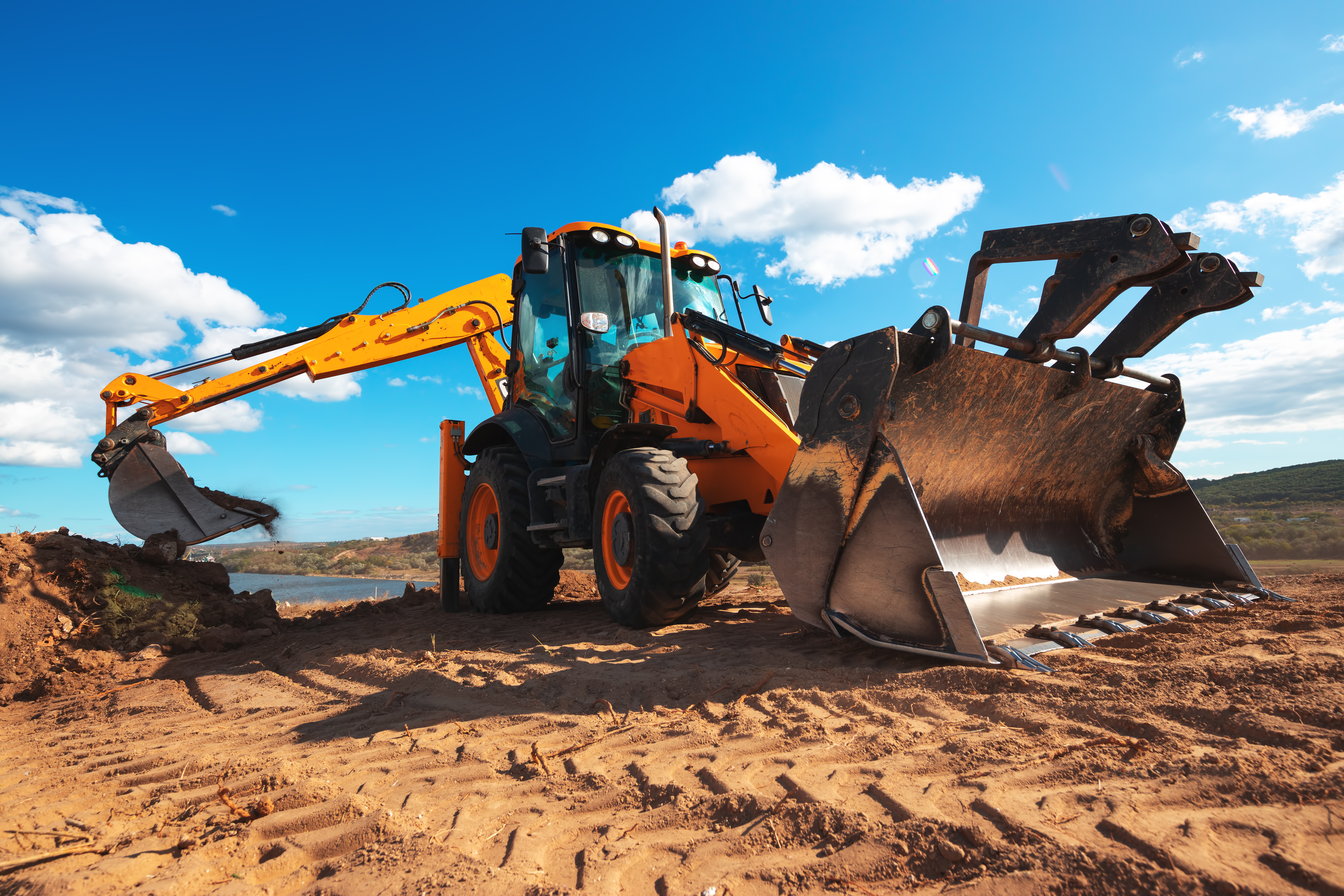 Orange and yellow backhoe loader working on sandy terrain with blue sky and clouds