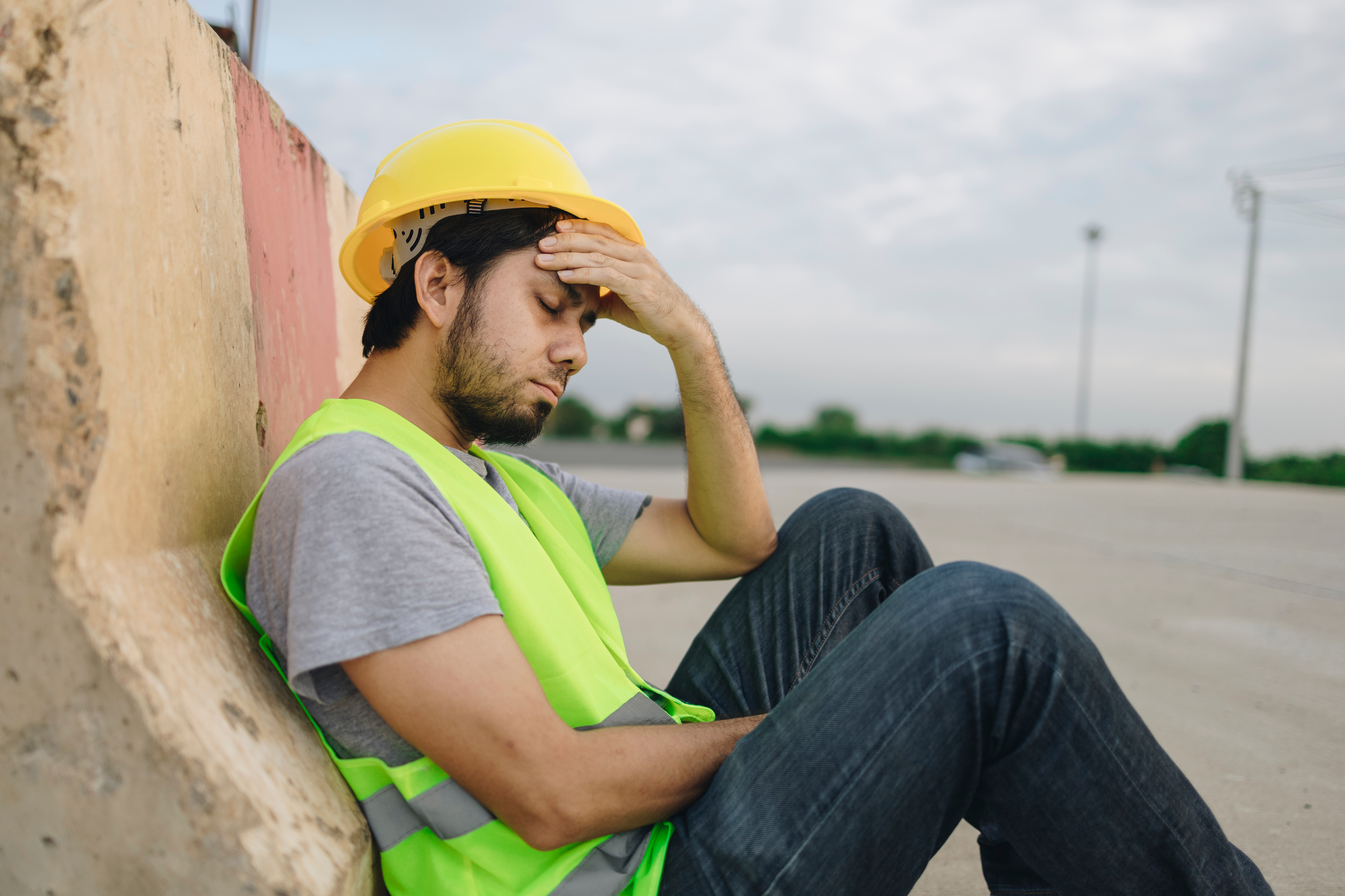 Construction worker in yellow hard hat and safety vest looking exhausted, leaning against wall