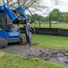 Blue tracked construction vehicle breaking concrete pavement near grassy field