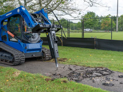 Blue tracked construction vehicle breaking concrete pavement near grassy field