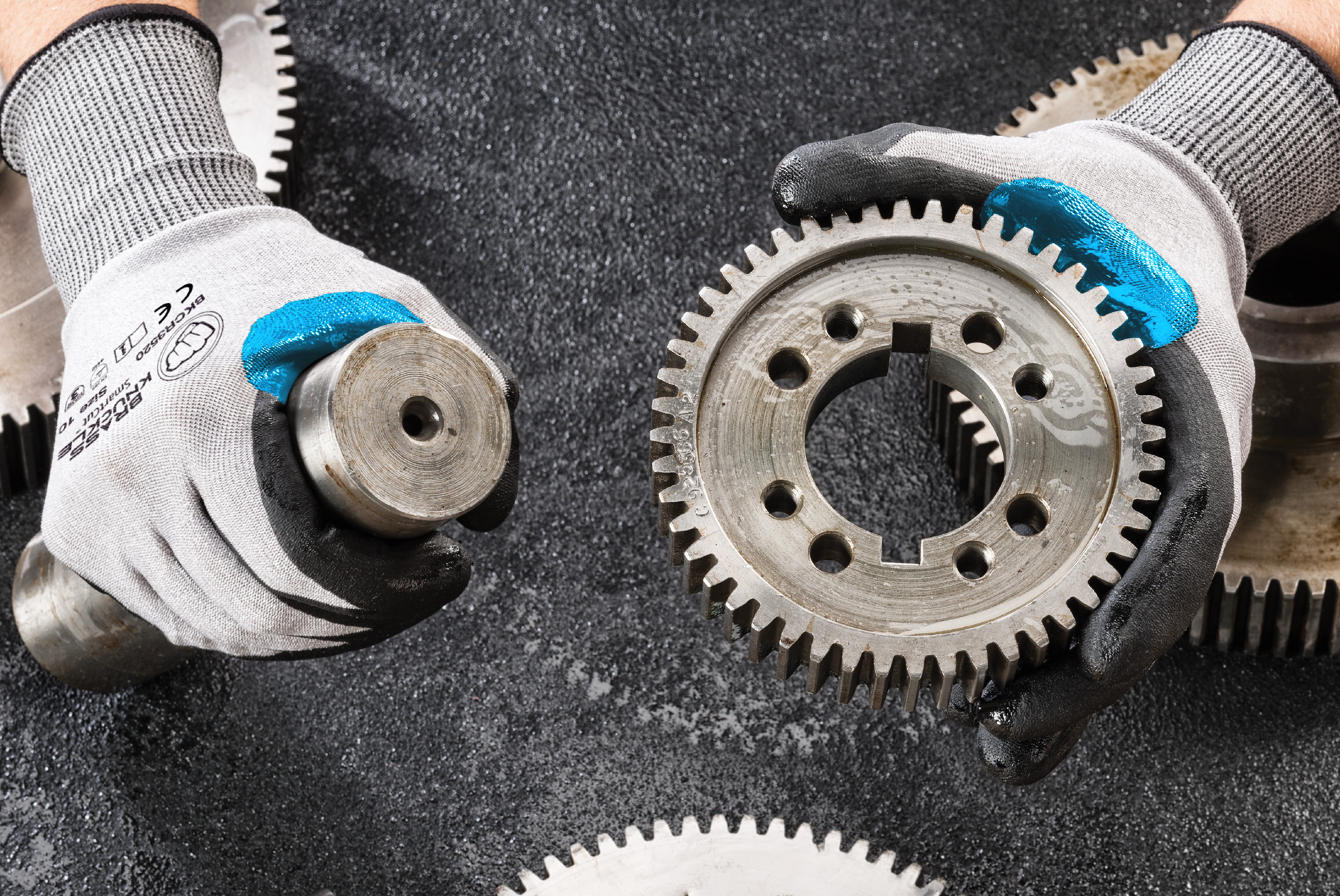 Industrial metal gears with blue-tipped work gloves on textured surface