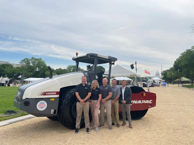 Four professionals stand beside a large white Dynapac construction roller on a grassy event area