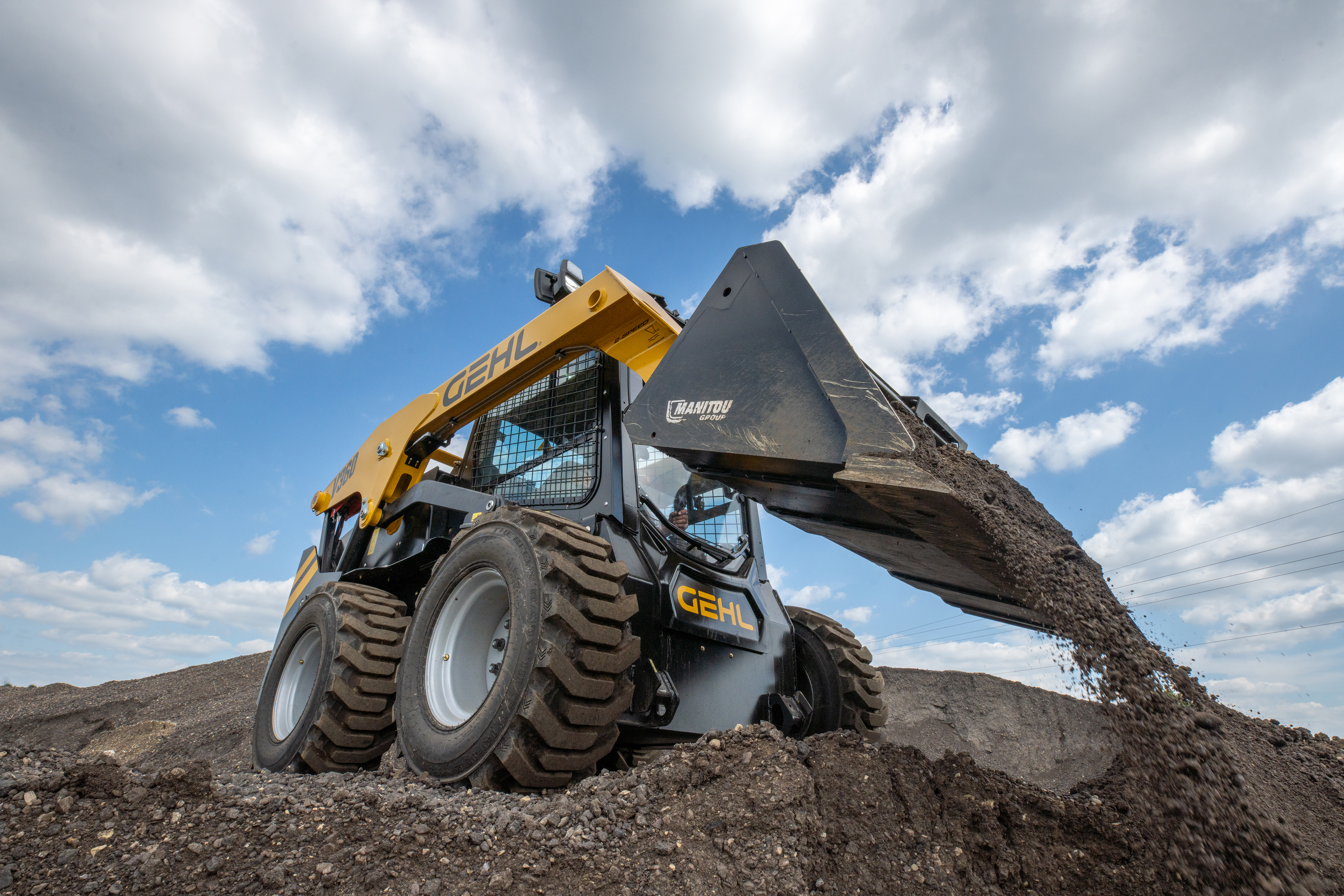 Yellow skid steer loader dumping dirt on rocky terrain with cloudy sky